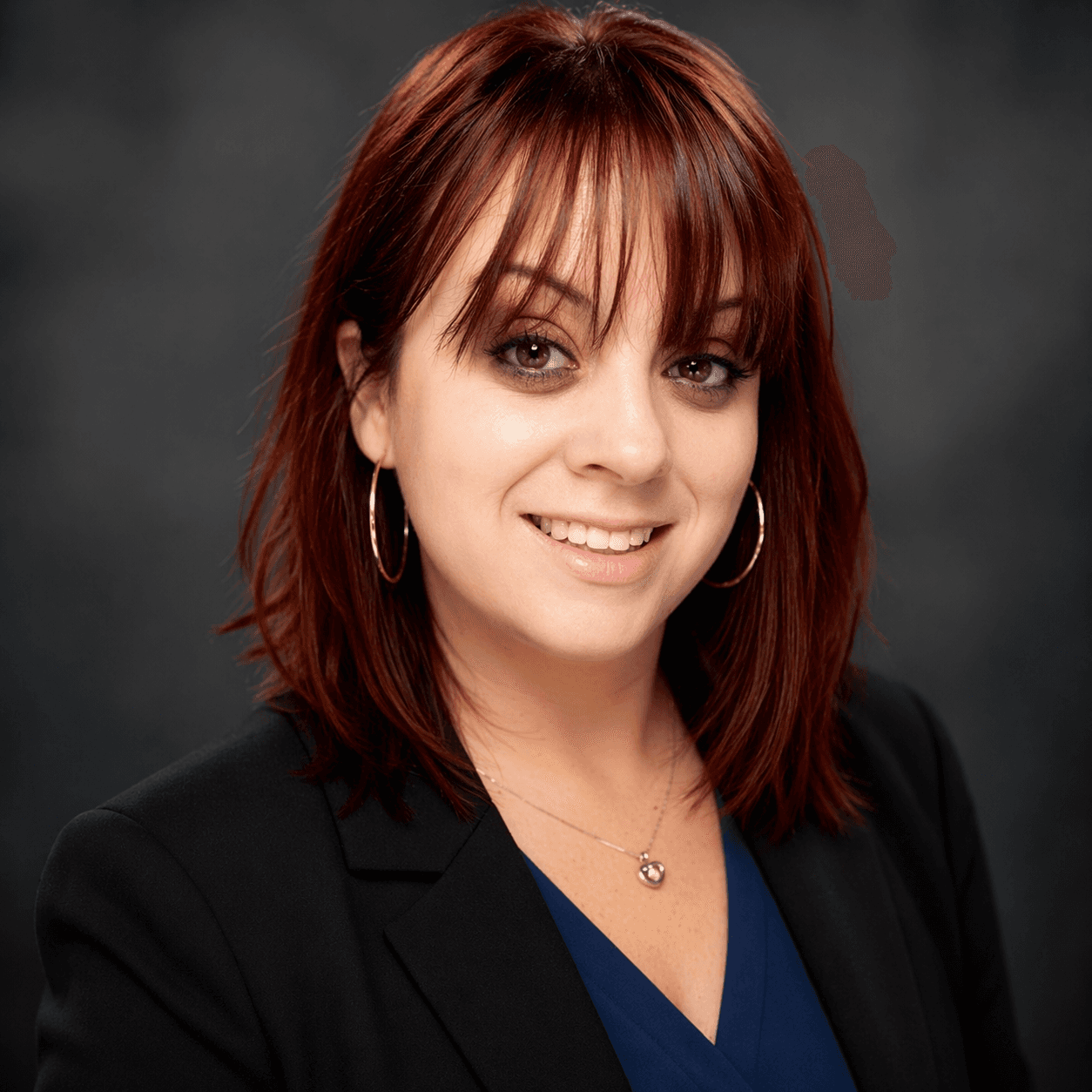 Smiling woman with auburn hair and bangs wearing a black blazer and gold hoop earrings.